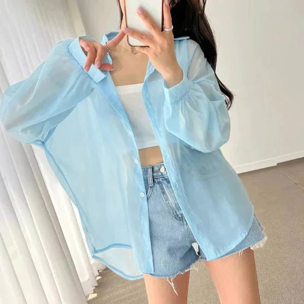 Woman wearing light blue sheer lightweight sun protection shirt over white crop top and denim shorts taking mirror selfie